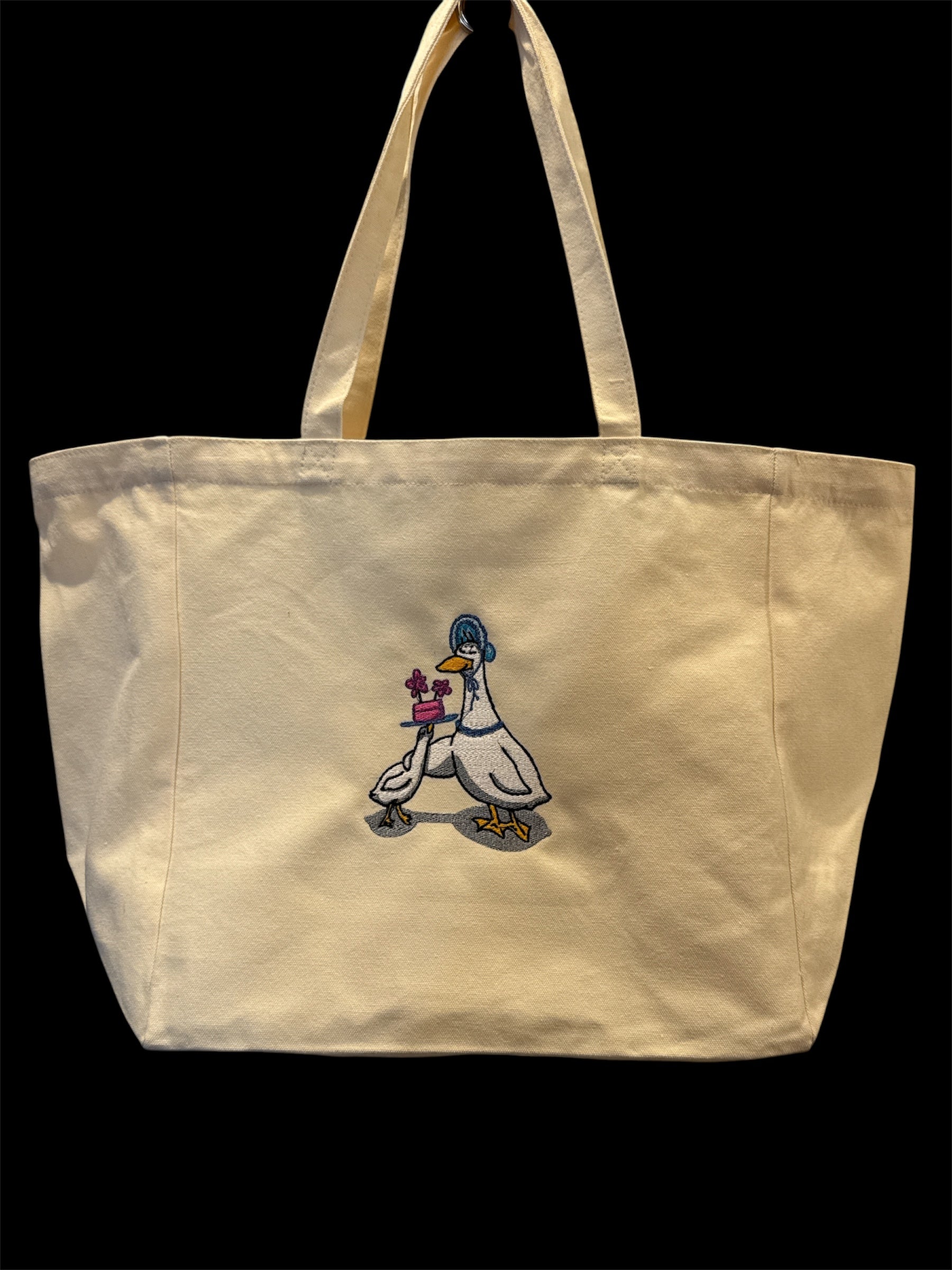Embroidered canvas tote bag depicting two cartoon geese, one wearing a blue bonnet and the other holding a small pink cake adorned with purple flowers